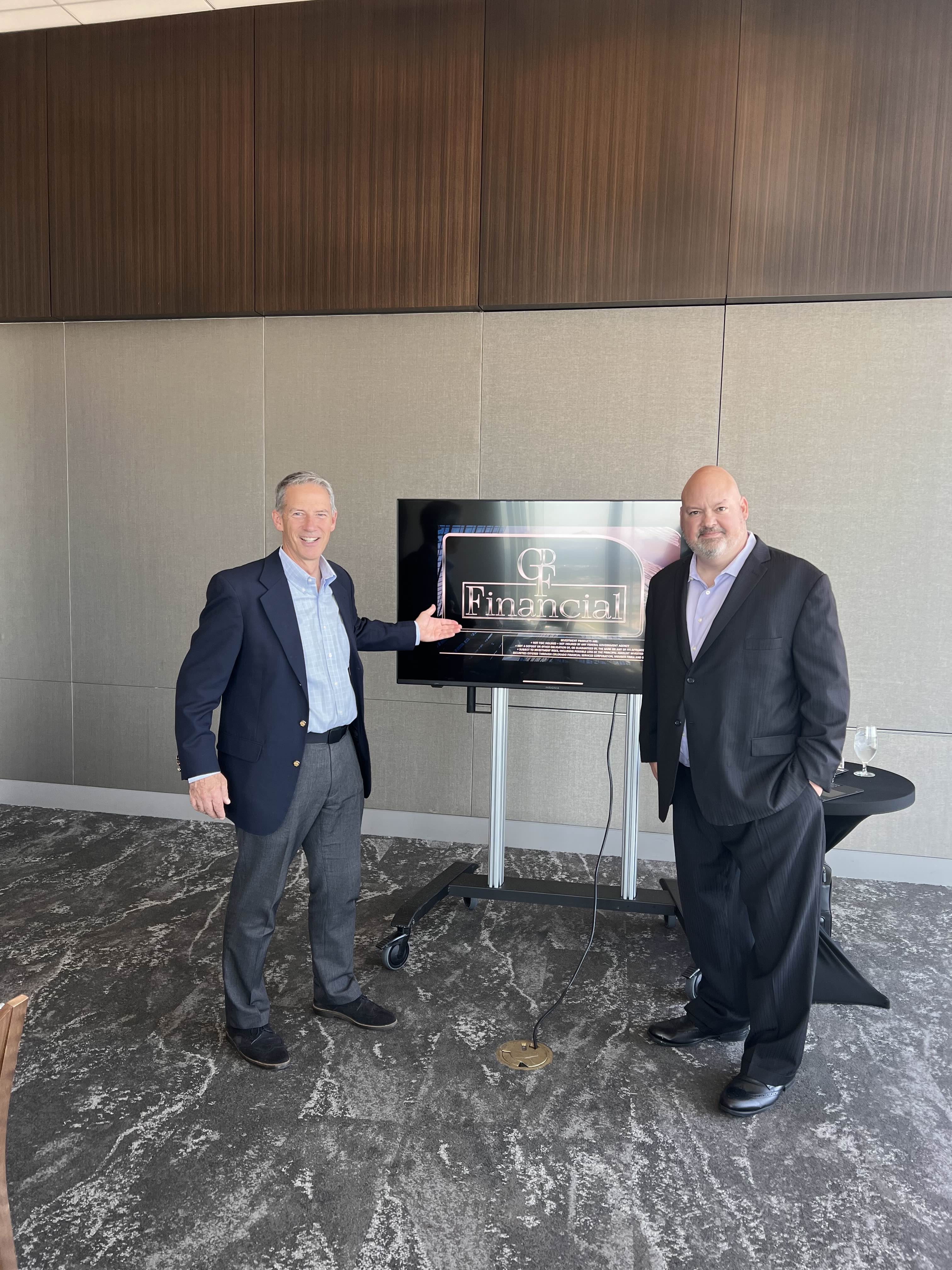 Two men in business attire standing on either side of a TV monitor displaying a logo reading 'GF Financial' in a conference room.