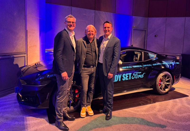 Three men in suits and casual clothes standing in front of a black Ford Mustang with READY SET Ford lettering.