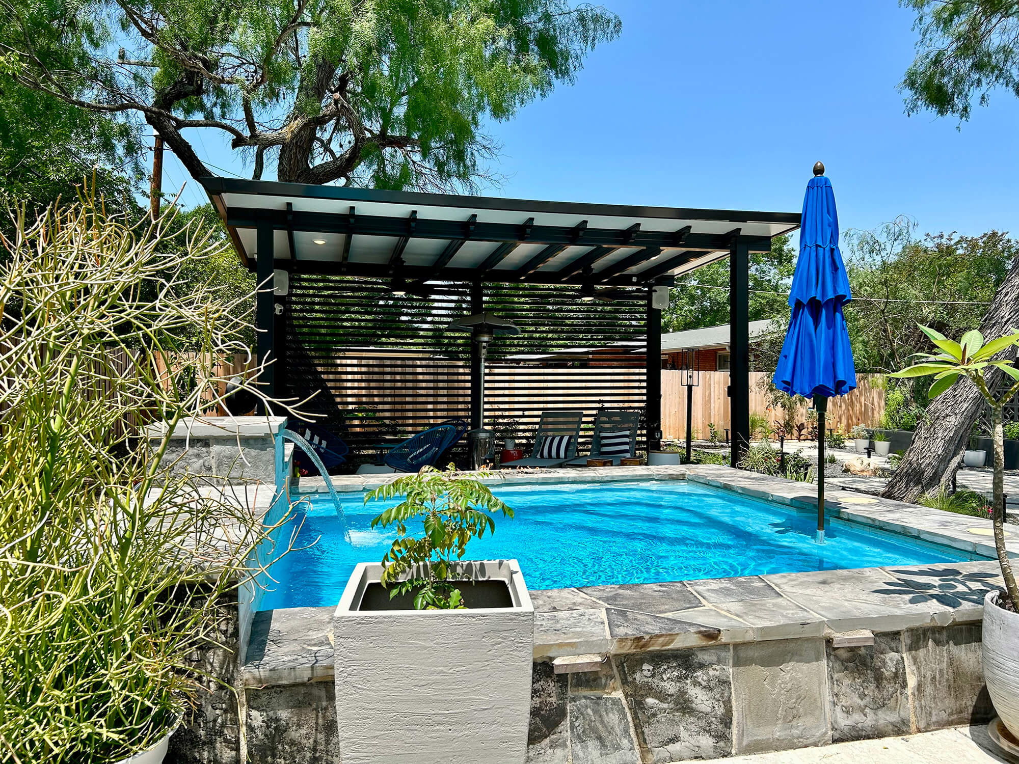 Backyard pool with pergola, blue umbrellas, and trees.