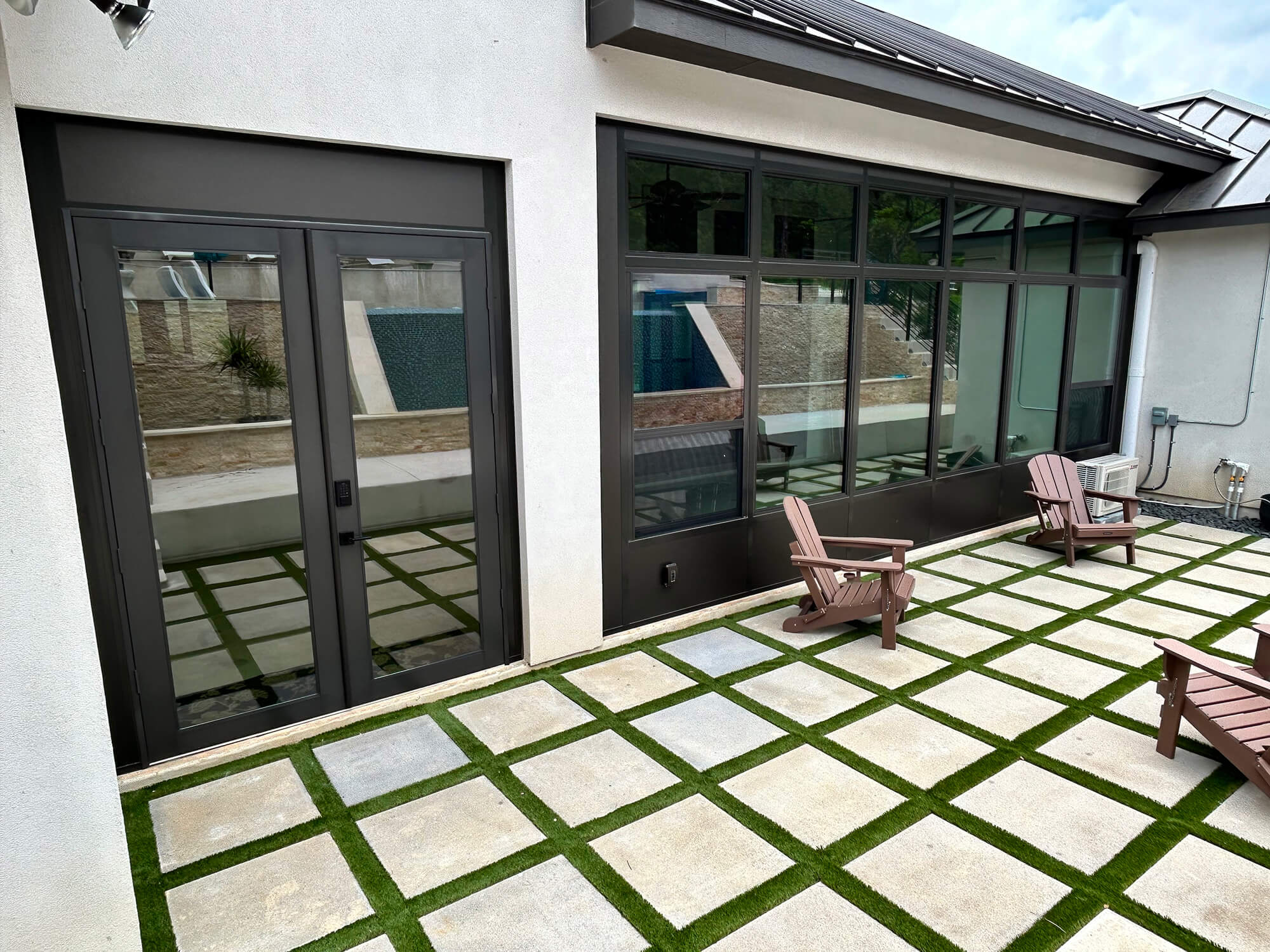 Modern patio with stone tiles and grass, wooden chairs, large windows and doors.