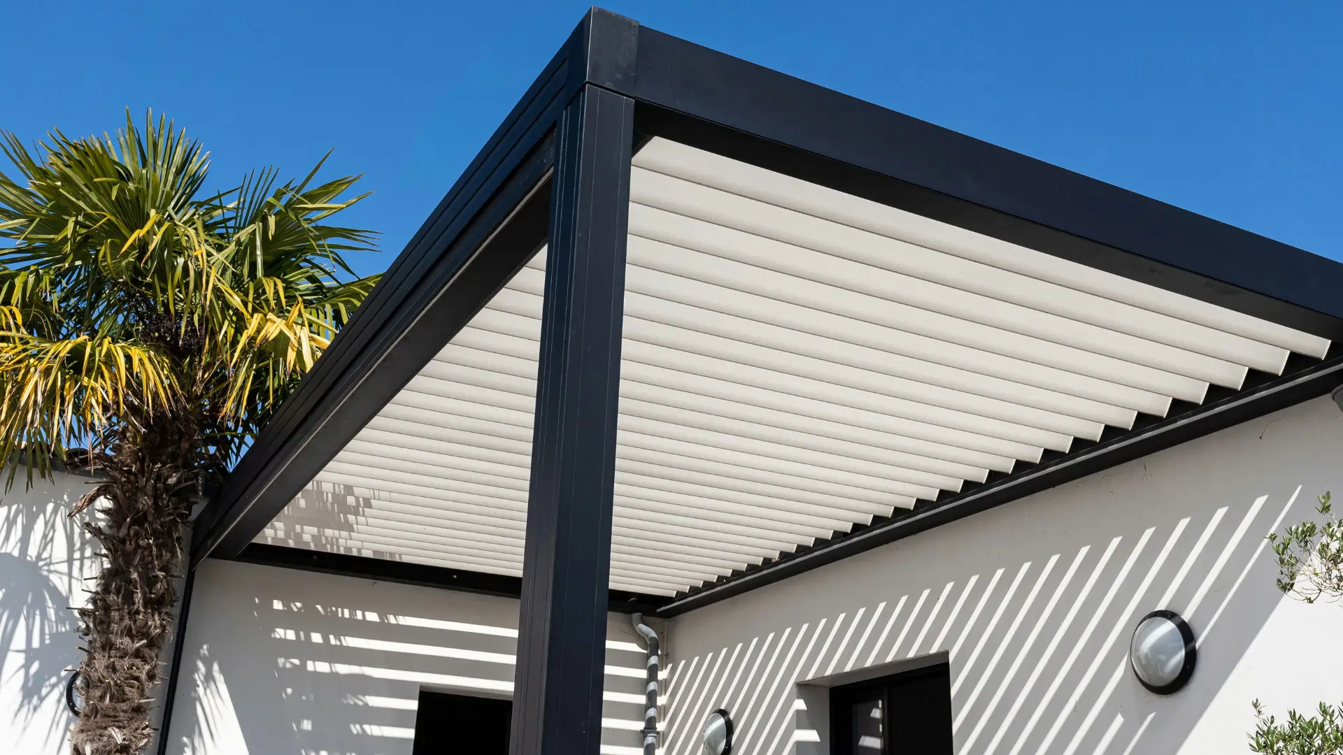 Modern pergola with black frame and white louvered roof casting striped shadows on white exterior wall under clear blue sky.