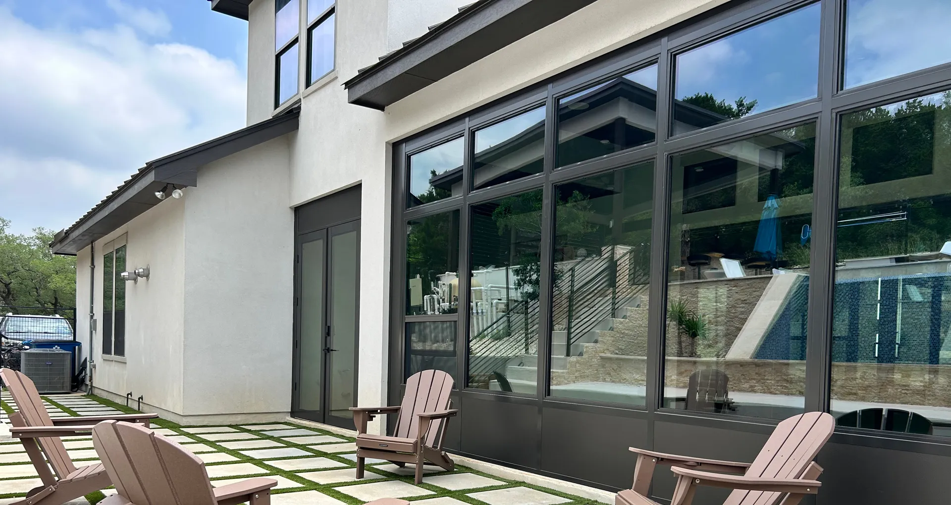Modern patio with brown Adirondack chairs on a tiled floor next to a house with large reflective windows.
