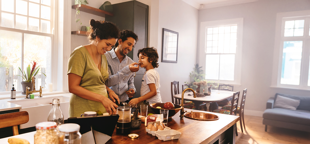 A mother, father, and young son cooking together in a sunny, open-plan kitchen.