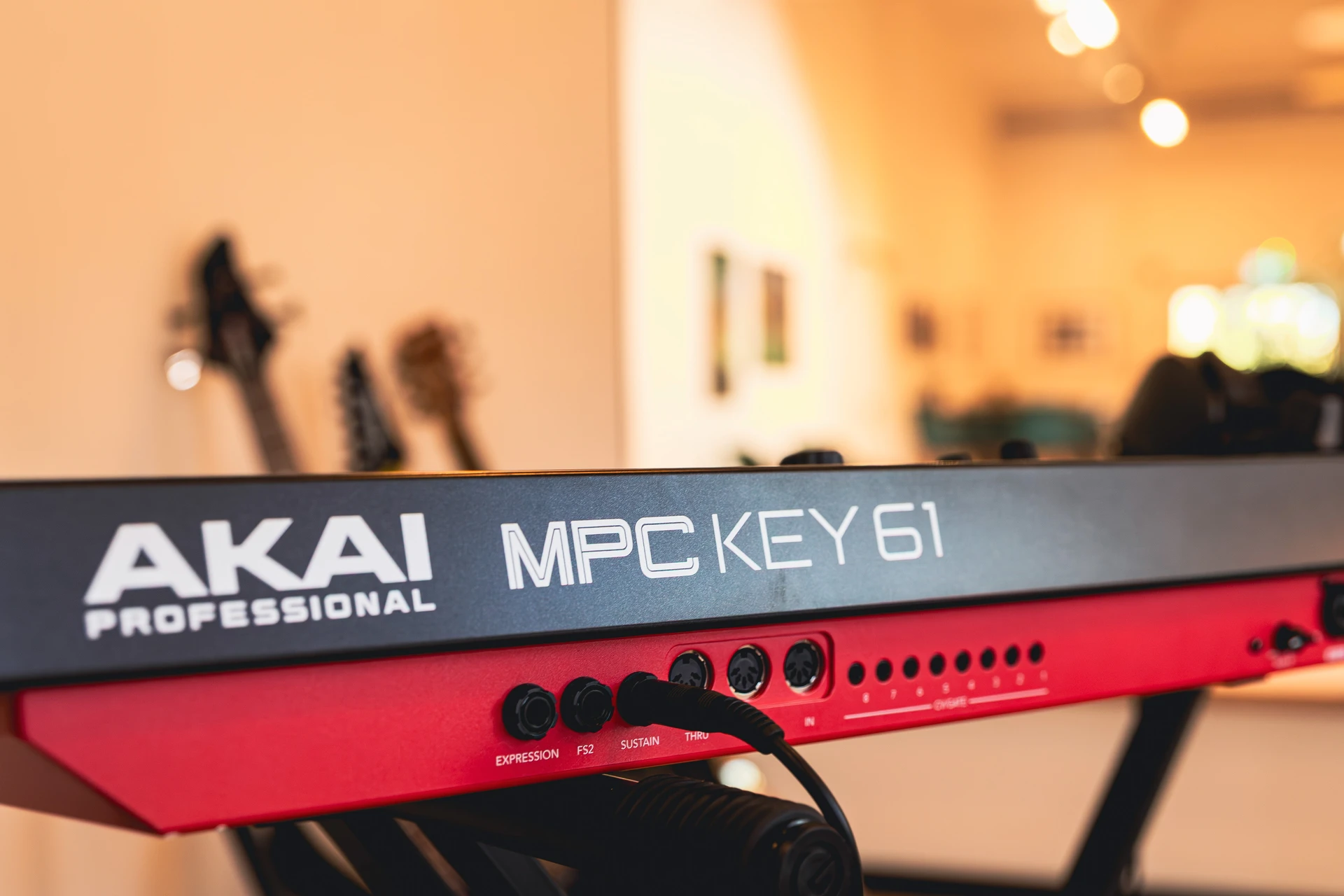 Rückansicht des roten AKAI Professional MPC Key 61 MIDI-Controllers mit angeschlossenen Kabeln vor verschwommenen Gitarren.