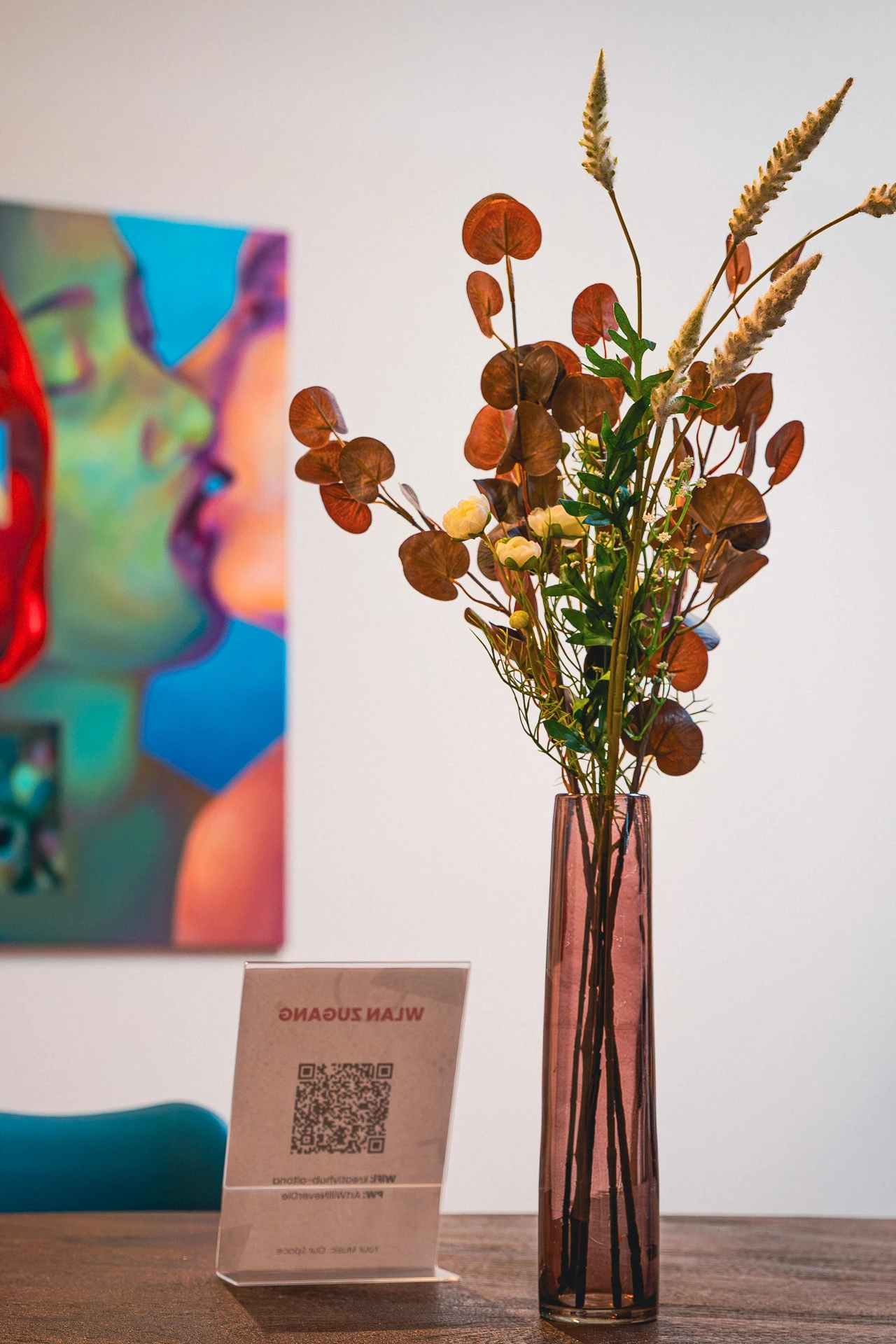 Vase mit getrockneten Blumen und Blättern auf einem Holztisch, im Hintergrund verschwommenes farbiges Gemälde an der Wand.