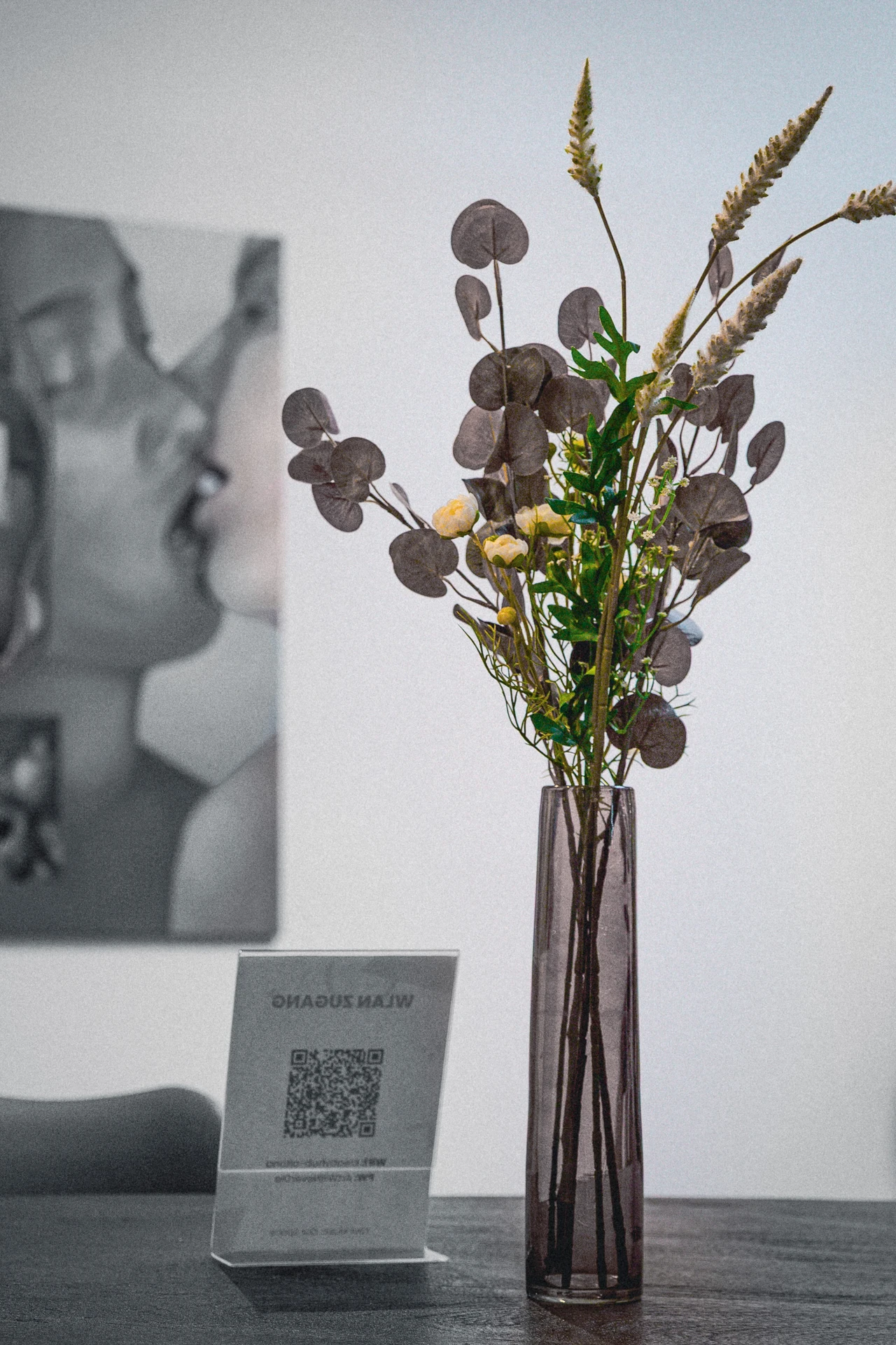 Vase mit getrockneten Blumen und Zweigen auf einem Tisch vor einem unscharfen Schwarz-Weiß-Foto von zwei sich küssenden Personen.