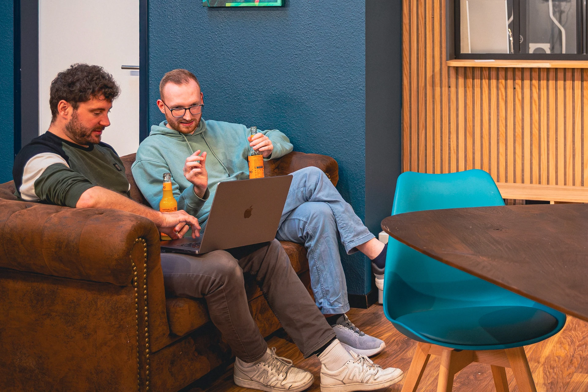 Zwei Männer sitzen auf einem braunen Sofa, schauen gemeinsam auf einen Laptop und halten jeweils eine Flasche mit orangem Getränk.