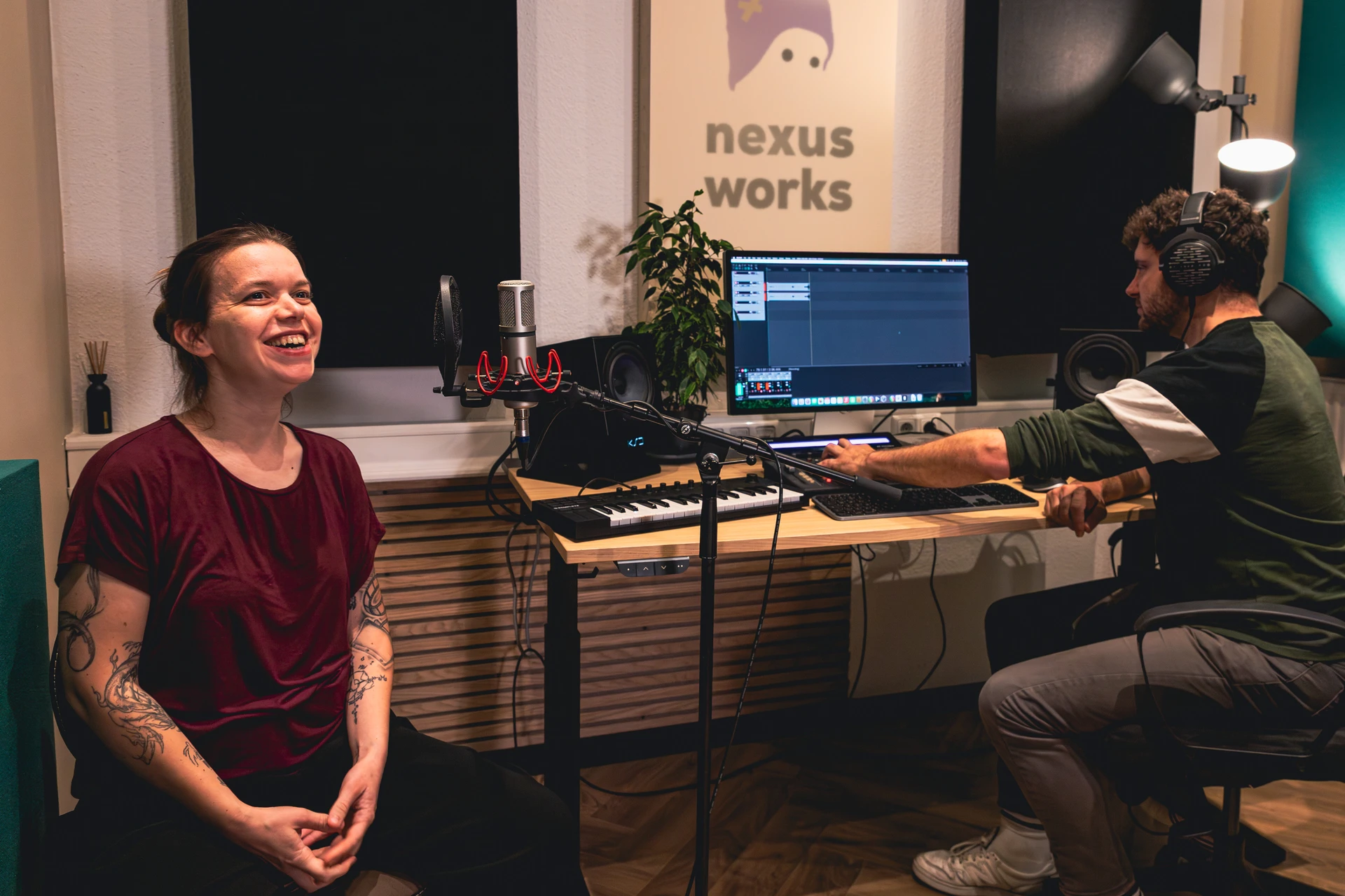 Frau mit Tätowierungen lächelt in einem Tonstudio vor einem Mikrofon, während ein Mann mit Kopfhörern an einem Computer mit Audiobearbeitungsprogramm arbeitet.
