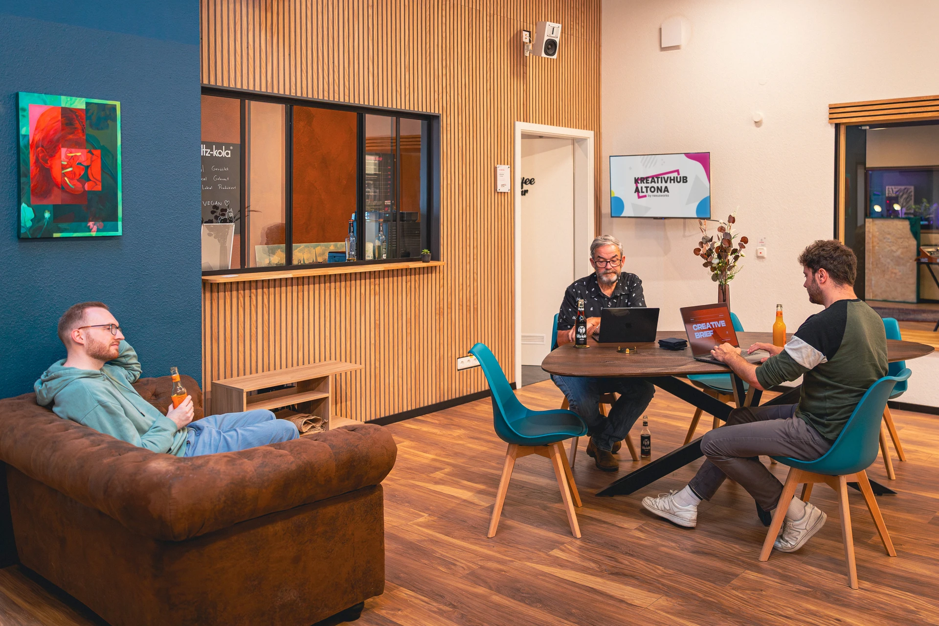 Drei Männer in einem modernen Gemeinschaftsarbeitsraum, zwei arbeiten an Laptops an einem Tisch, einer sitzt entspannt auf einem braunen Sofa und hält eine Flasche.