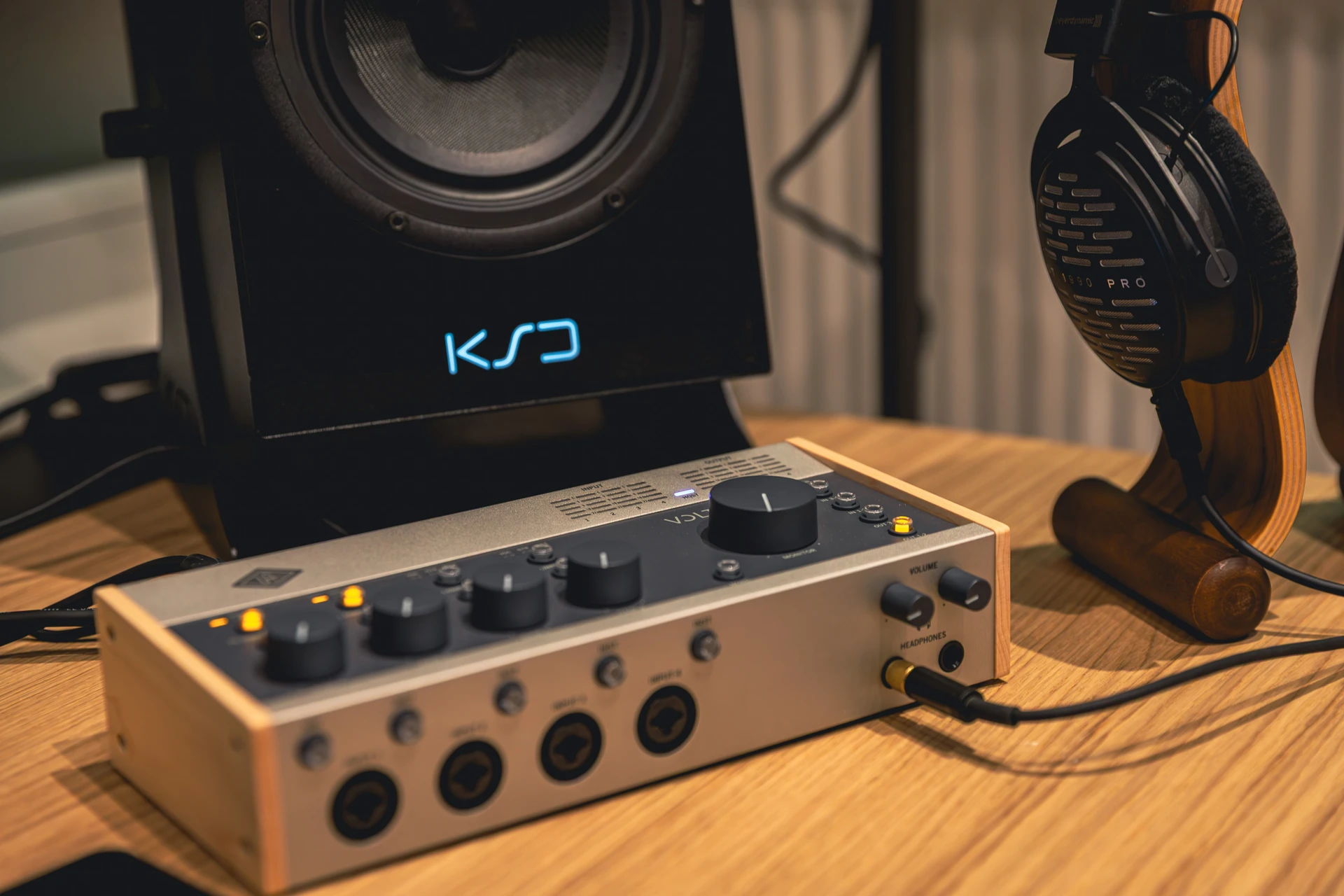 Audiogerät mit Drehreglern und Anschlüssen auf einem Holztisch, daneben ein Hörer auf einem Ständer und ein Lautsprecher mit blau beleuchtetem Logo.