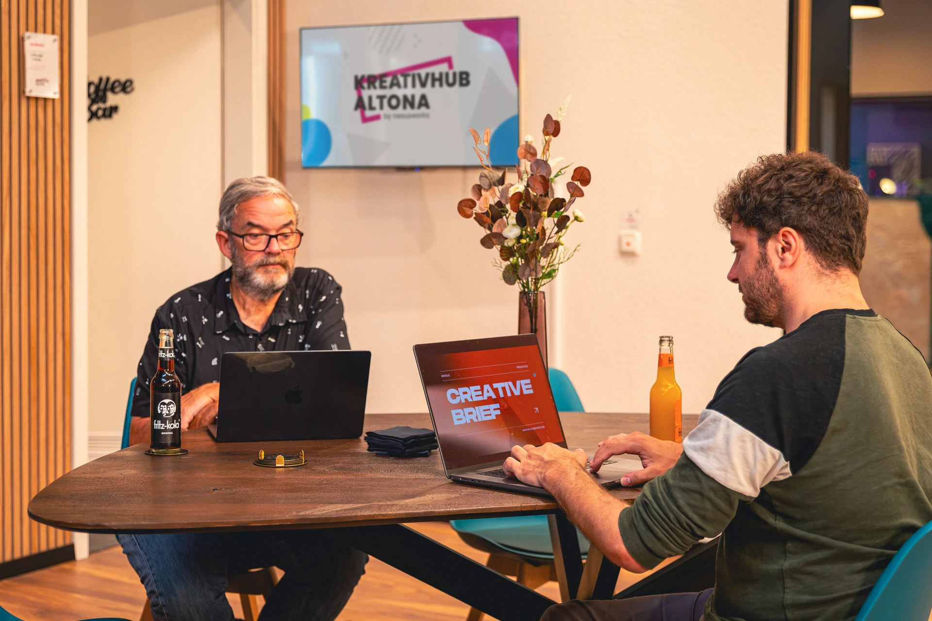 Zwei Männer arbeiten an Laptops an einem Holztisch in einem modernen Büro mit Bildschirm 'KREATIVHUB ALTONA' im Hintergrund.