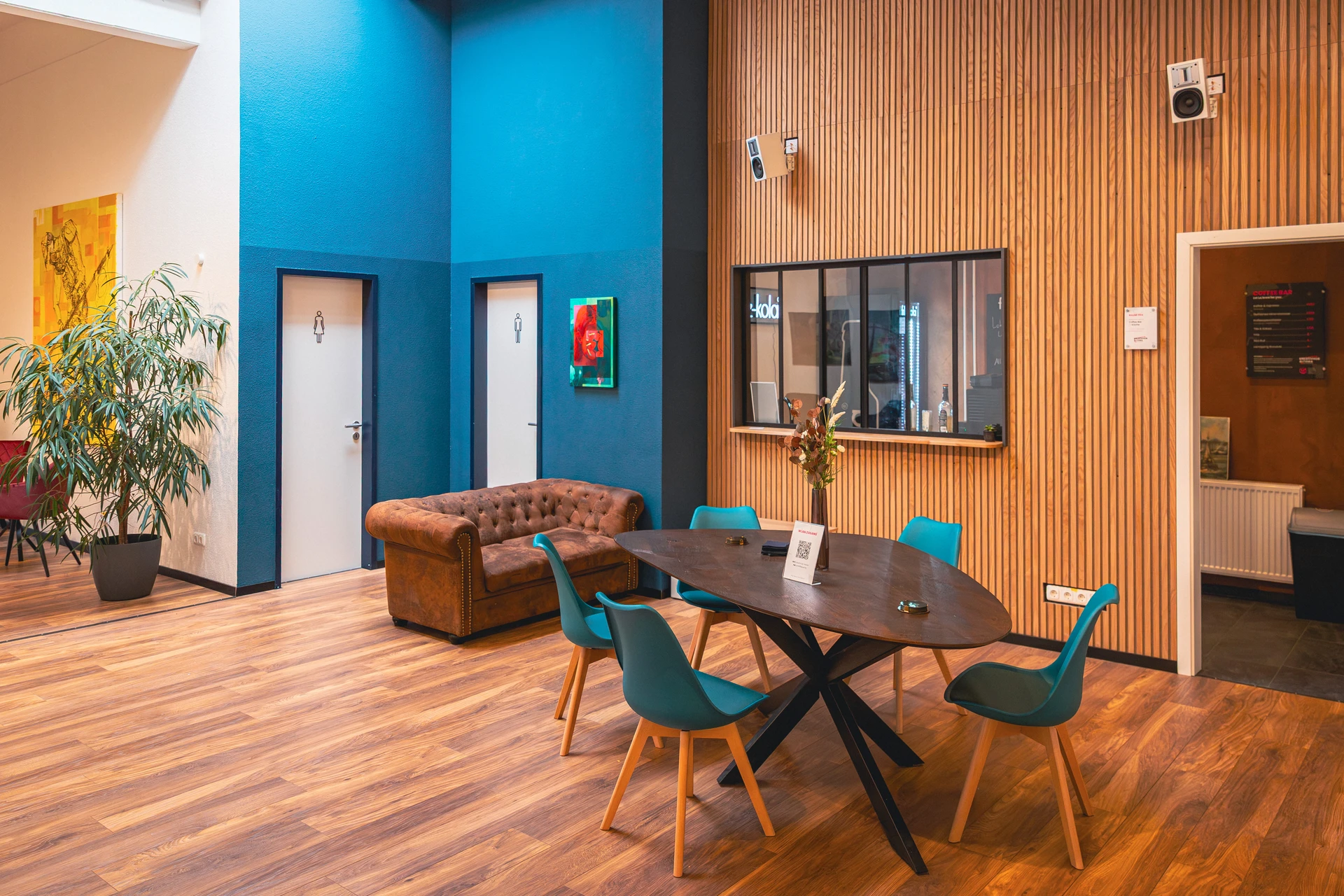 Moderner Aufenthaltsraum mit Holzboden, braunem Sofa, blau gestrichenen Wänden, blau-grünen Stühlen um einen Holztisch und zwei Türen für Damen- und Herrentoilette.