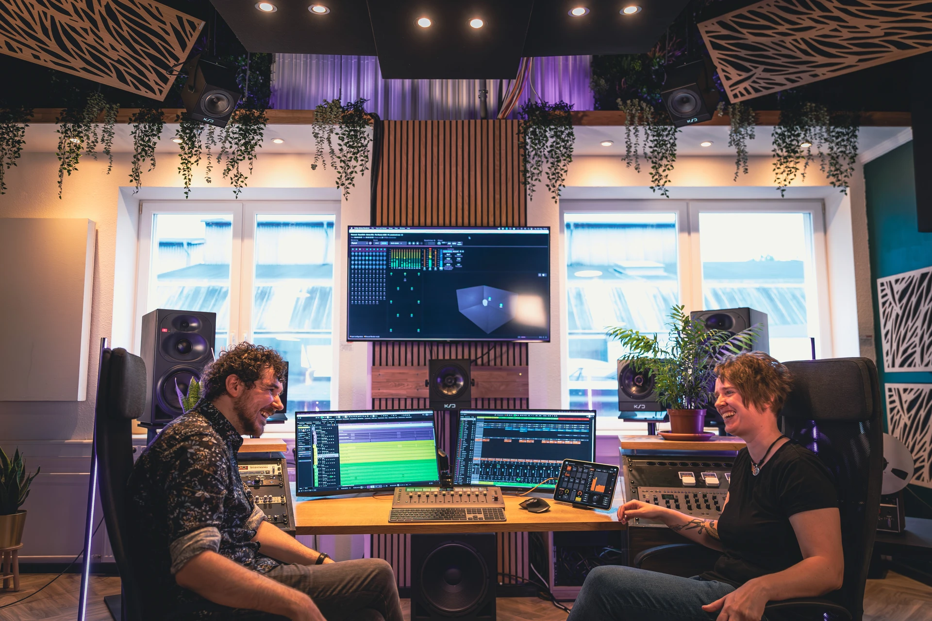 Zwei Menschen lächeln sich gegenüber sitzend in einem modernen Musikstudio mit mehreren Bildschirmen, Lautsprechern und Pflanzen.