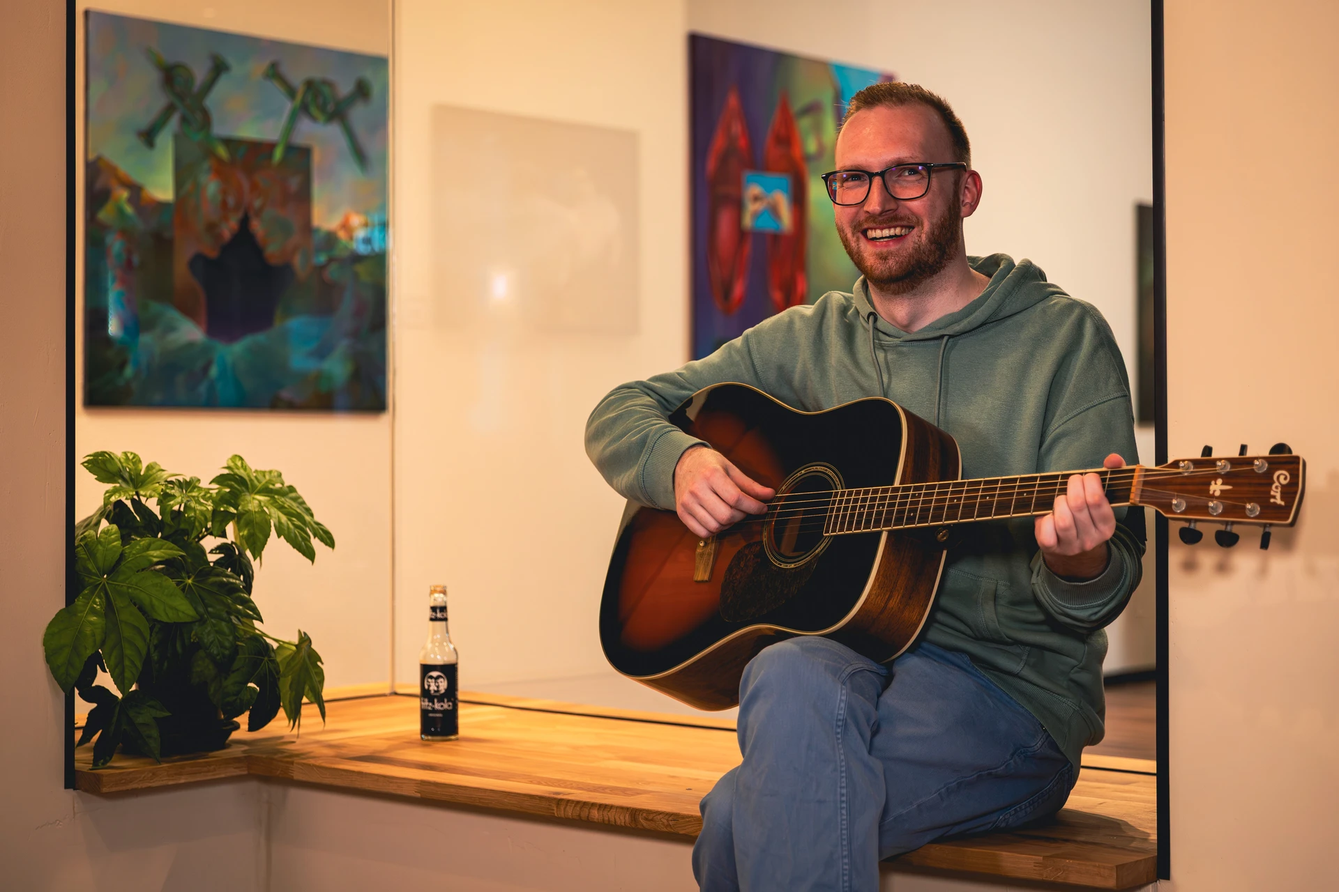 Mann mit Brille und grünem Kapuzenpullover sitzt lächelnd auf einer Holzbank und spielt akustische Gitarre in einem gemütlichen Raum mit Pflanzen und Wandbildern.