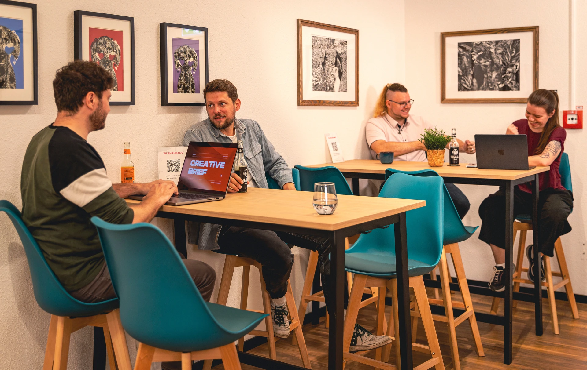 Vier Personen sitzen an zwei Tischen in einem modernen Arbeitsbereich, zwei arbeiten am Laptop, die anderen unterhalten sich bei Getränken.