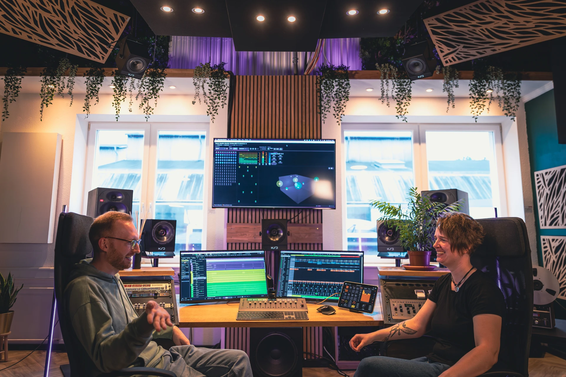 Zwei Personen unterhalten sich in einem modernen Tonstudio mit mehreren Monitoren, Lautsprechern und Pflanzen.