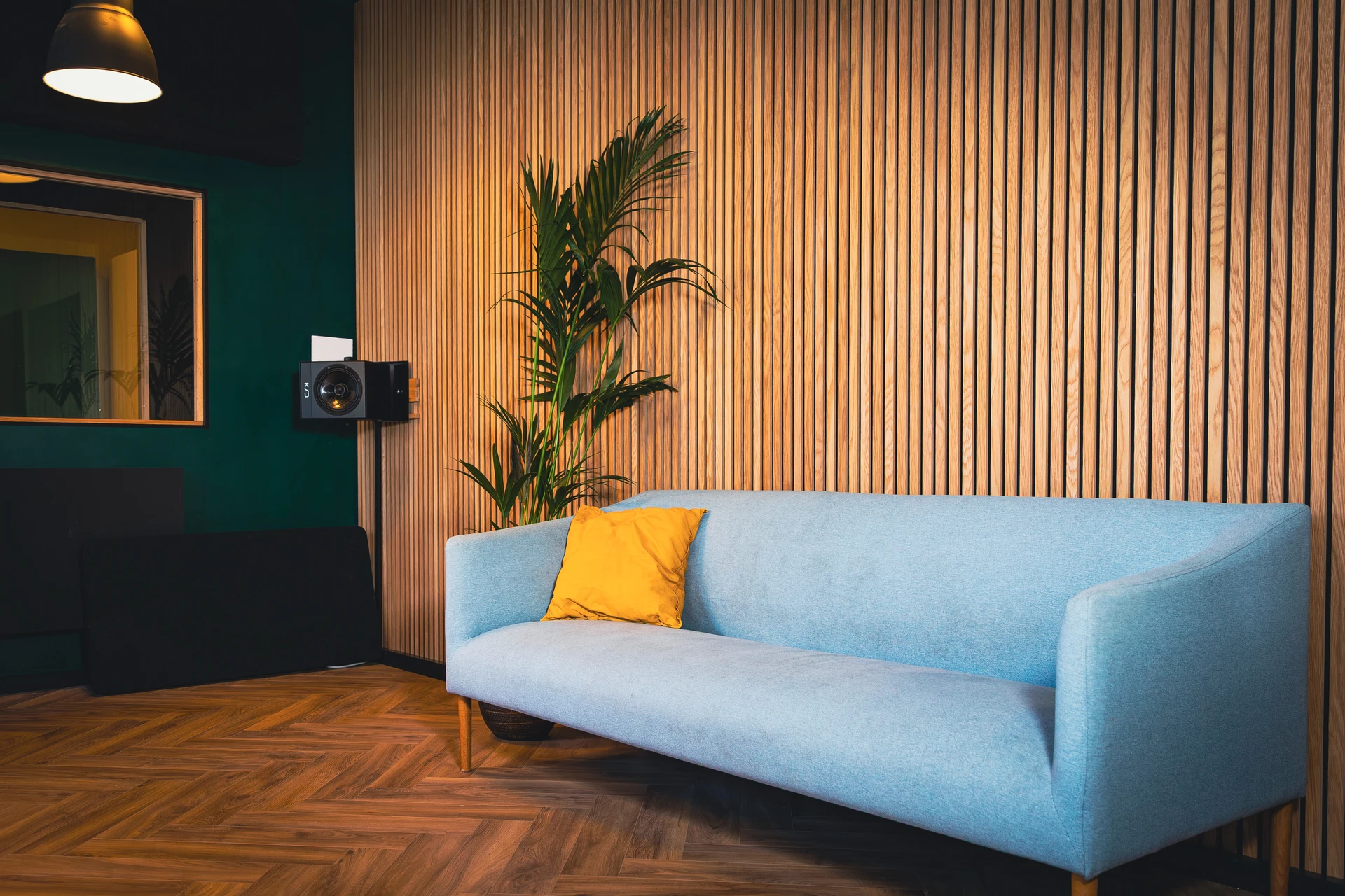 Hellblaues Sofa mit orangem Kissen vor einer Holzlattenwand, daneben eine grüne Pflanze und eine schwarze Wandlautsprecherbox.