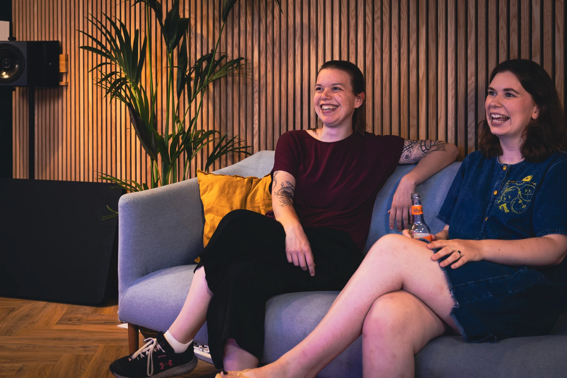 Zwei lächelnde Frauen sitzen auf einem grauen Sofa vor einer Holzlattenwand, eine hält eine Flasche Getränk.
