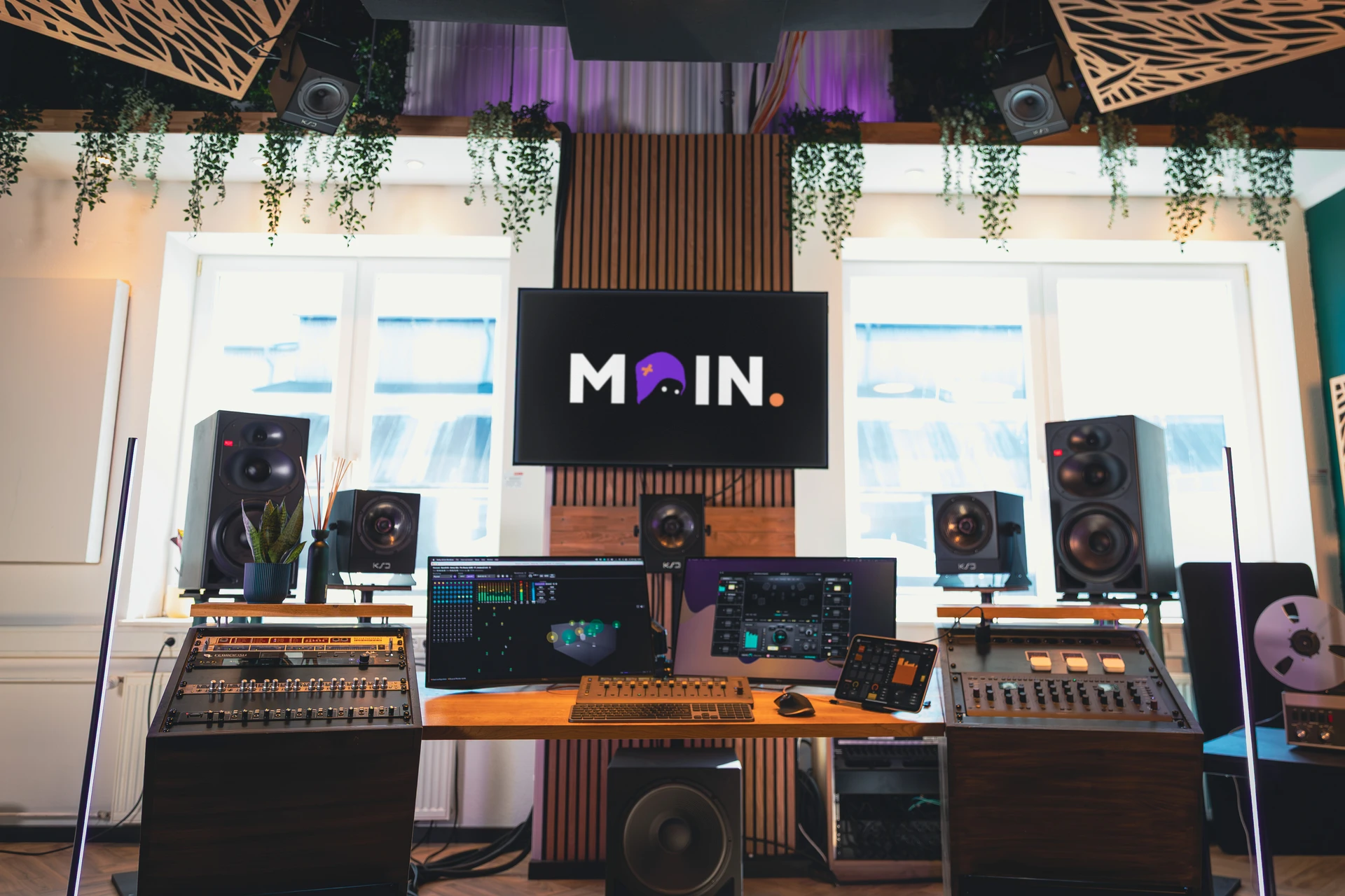 Moderne Musikproduktionsstation mit zwei Monitoren, mehreren Lautsprechern, Mischpulten und Pflanzen vor zwei hellen Fenstern.