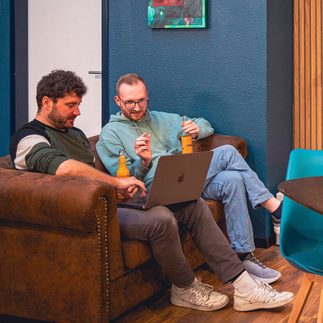 Zwei Männer sitzen auf einem braunen Sofa, schauen gemeinsam auf einen Laptop und halten jeweils eine Flasche Getränk in der Hand.