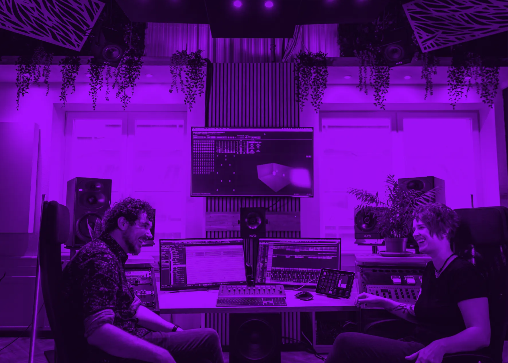 Zwei Personen sitzen lachend an einem Tisch mit Audio-Equipment und Monitoren in einem Studio.