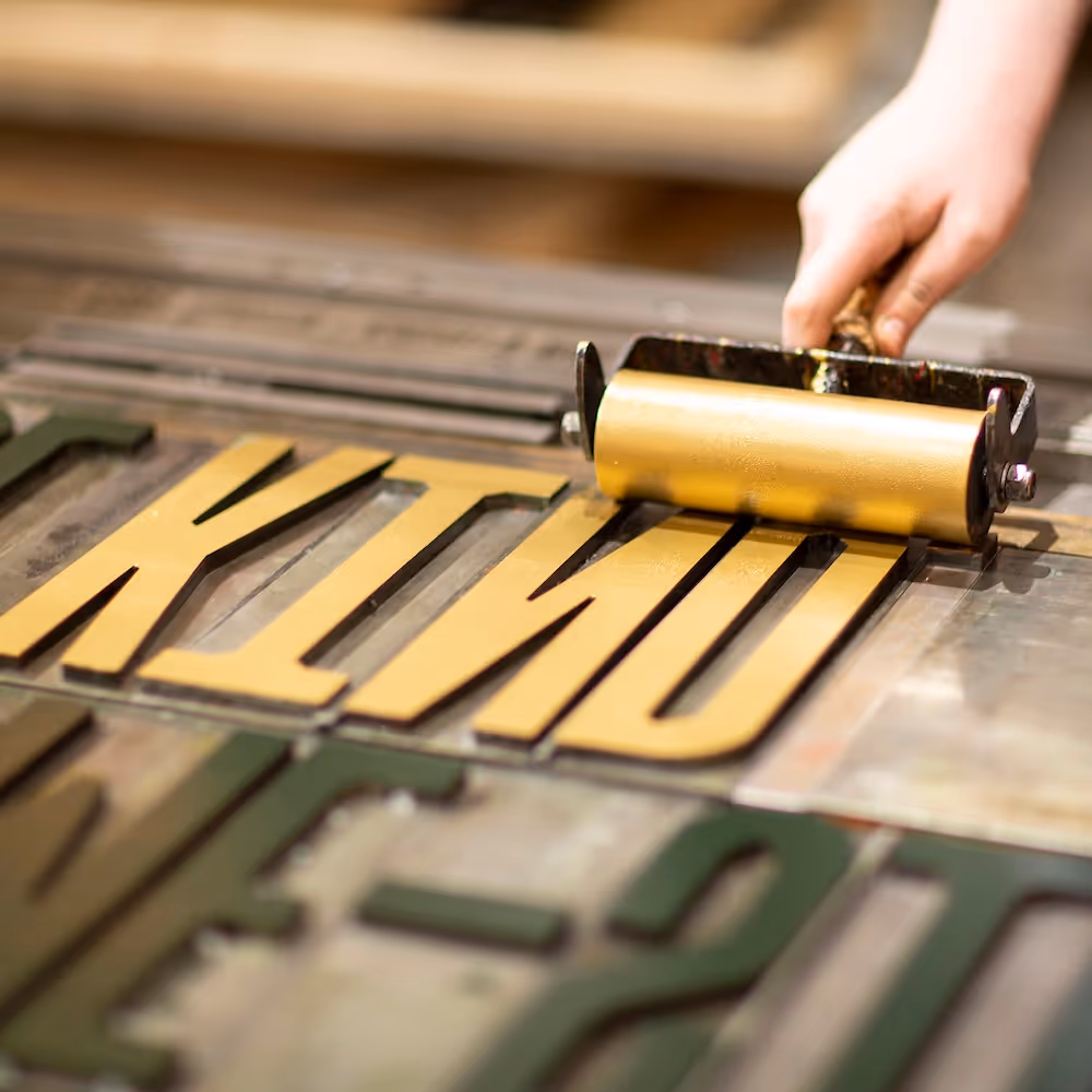 A workshop participants rolls gold ink onto the steel letters of typeface GRAFT, spelling the word KIND.