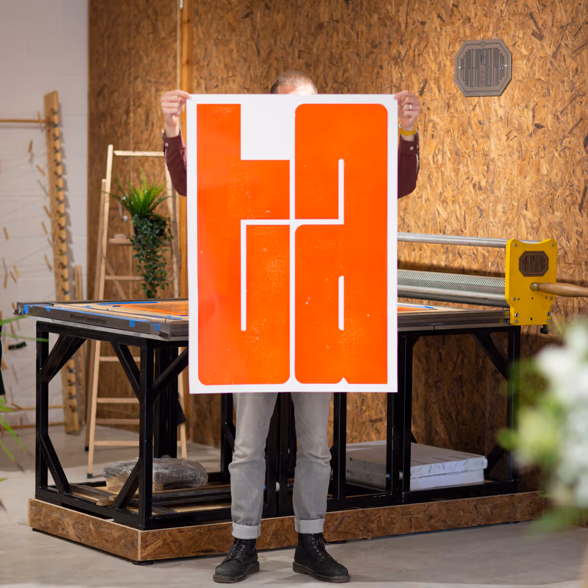 A large print featuring the word 'TA' in bright orange ink, being held up in front of the People Powered Press by Oli Bentley, who designed and made the print