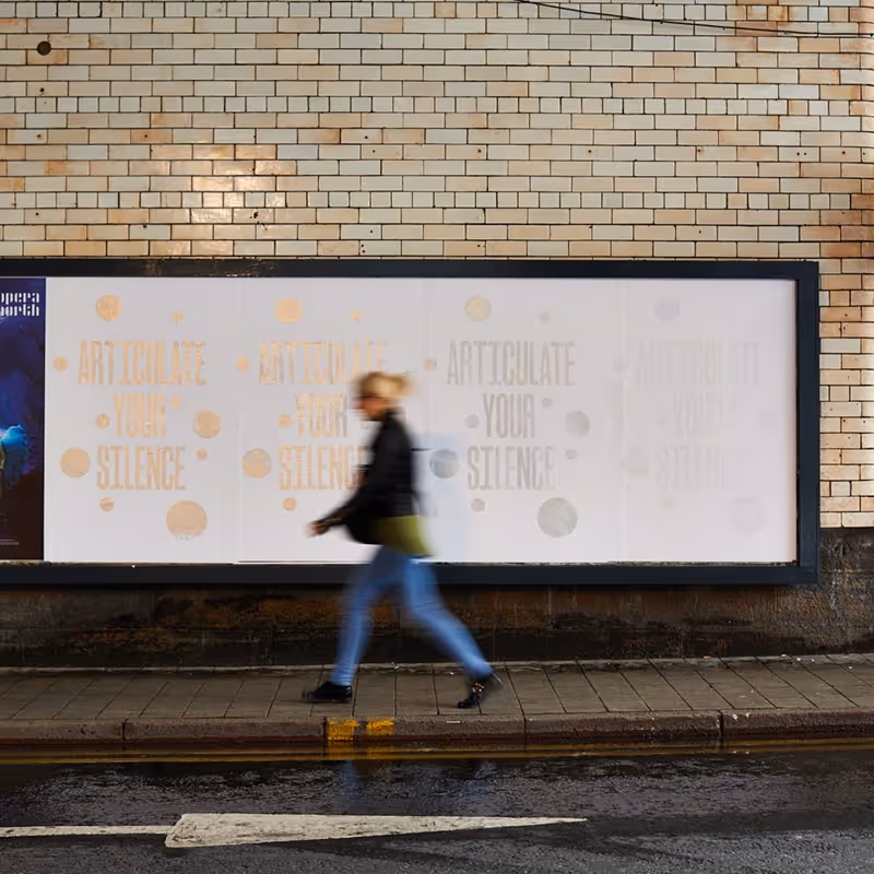 Four large murals pasted to a boarding underneath a railway bridge and beside a road. A woman walks in front of them, motion-blurred. The murals all read, 'ARTICULATE YOUR SILENCE' in metallic ink; from left to right they appear progressively more faded.