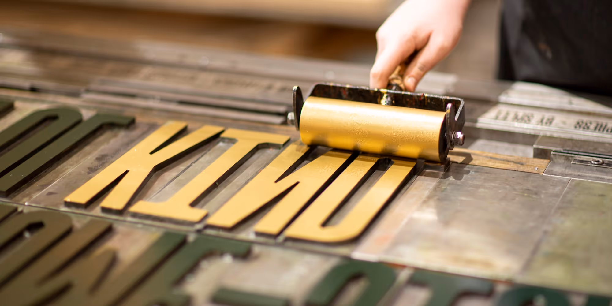 A hand is visible, holding a roller covered in gold ink and covering the surface of steel letters in the print-bed of the People Powered Press. The word reads, 'KIND'.