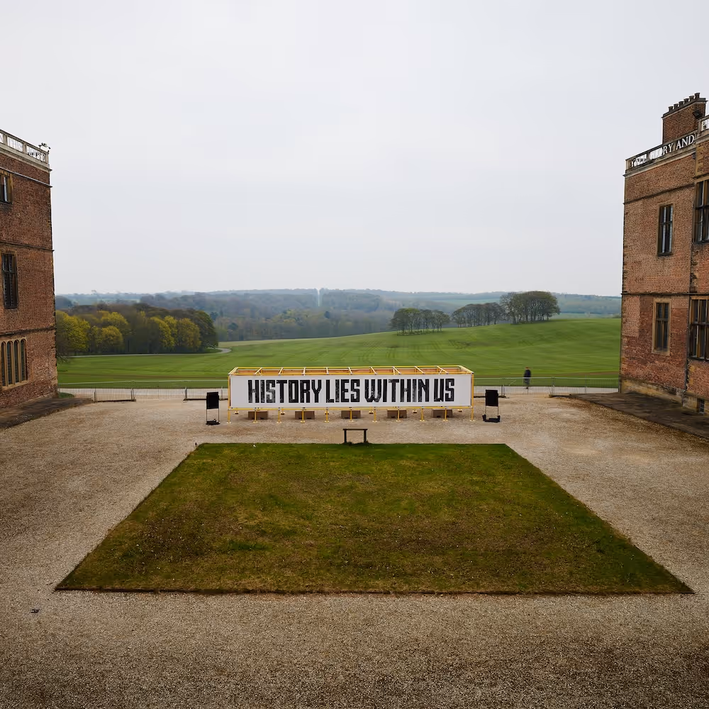 A giant mural reading 'History Lies Within Us' pasted to a wide yellow scaffolding structure, standing in the courtyard of a stately home. Hills and woods stretch into the distance.