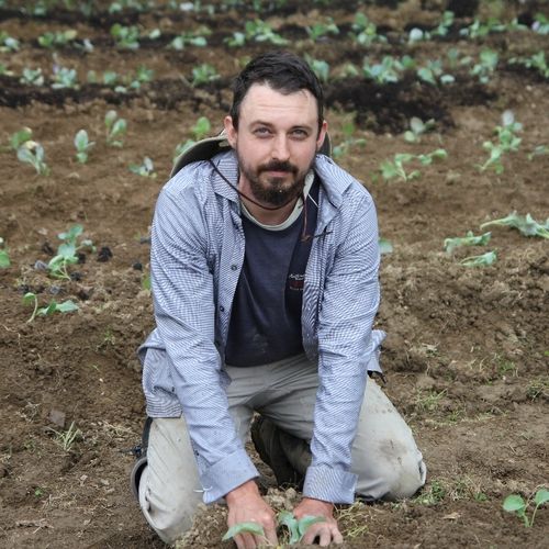 A picture of Matt kneeling in a field of seedlings. He is wearing khakis, a dark blue t-shirt, and a light blue long sleeve button up.