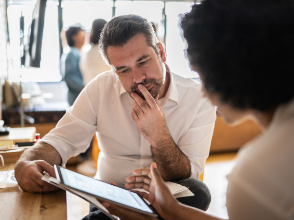 Two people in a discussion, one holding a tablet and the other thoughtfully looking at it.