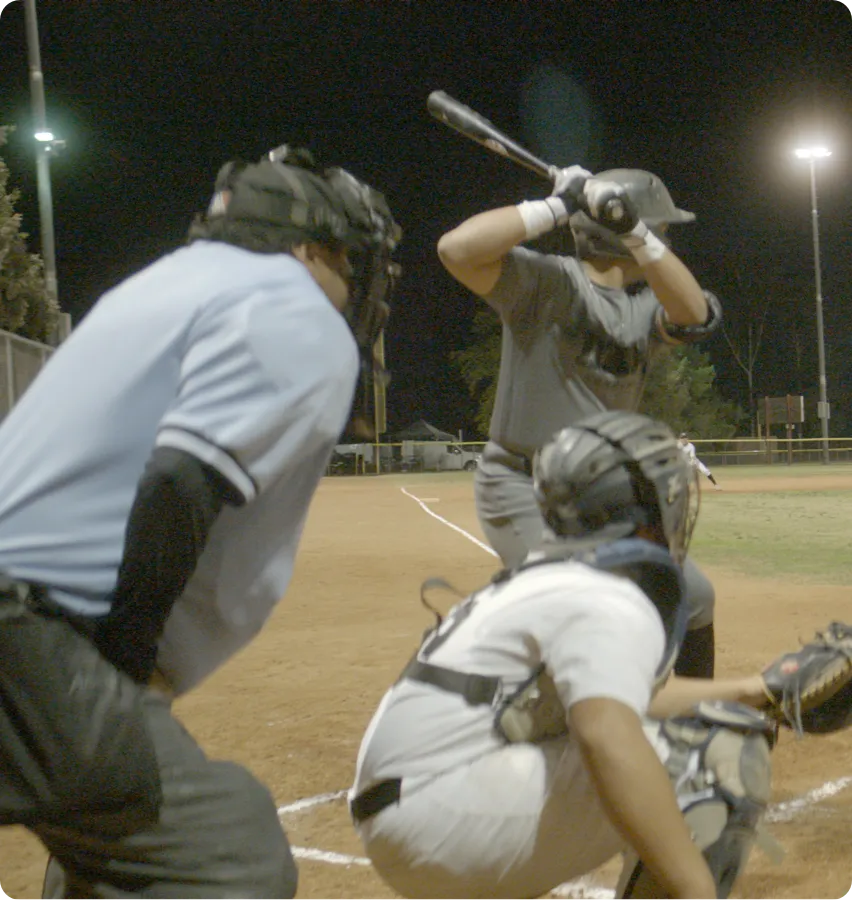 Nighttime baseball scene with batter preparing to swing, catcher crouched behind home plate, and umpire observing closely.