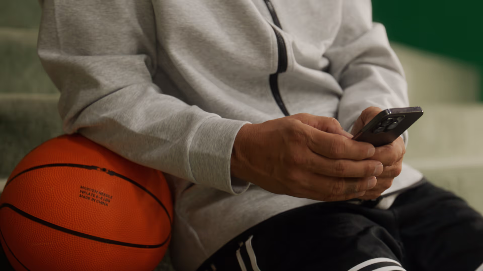 Person in gray hoodie holding a smartphone with an orange basketball resting against their side.