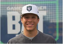 Smiling young man wearing a gray shirt and a white cap with a black logo standing in front of a mesh backdrop.