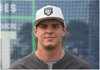Young man wearing a gray and white baseball cap with a 'BAT' logo, standing outdoors in front of a blurred city background.