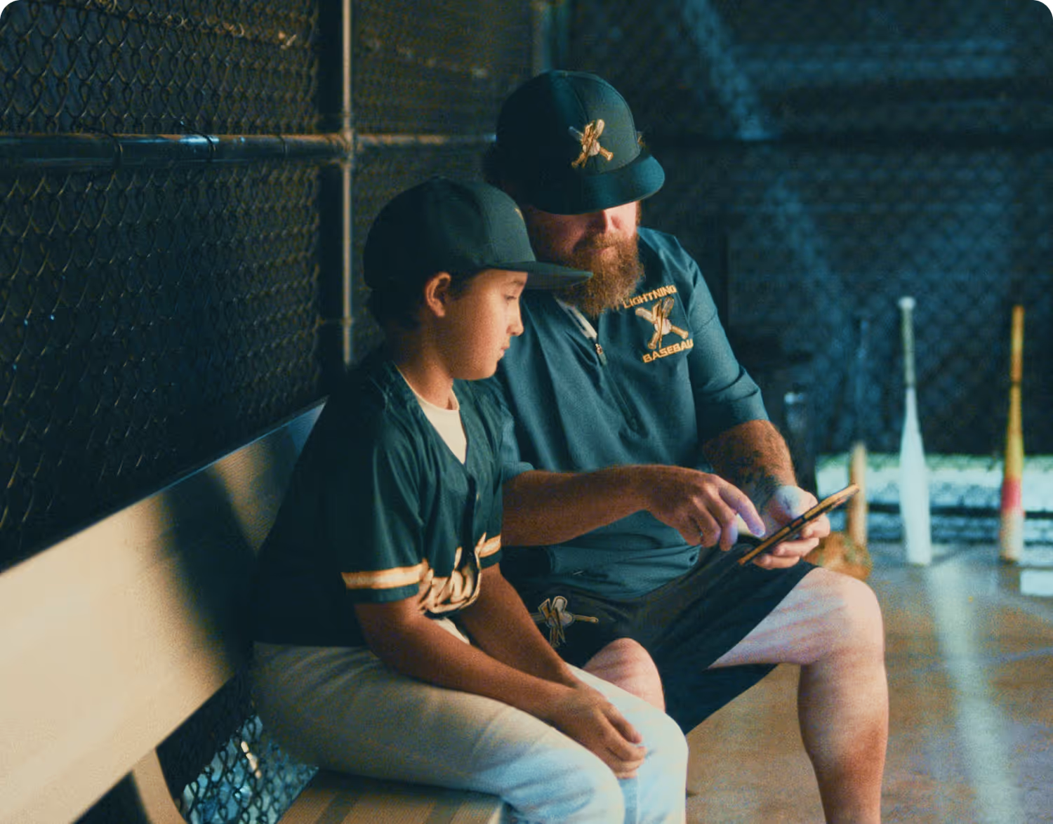 Baseball coach showing something on a tablet to a young player sitting on a bench in a dugout.
