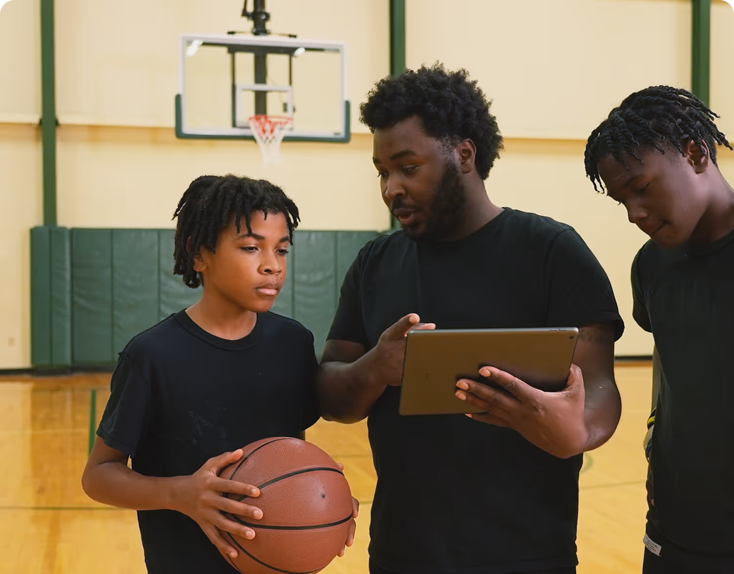 Basketball coach discussing strategy on a tablet with two young players in a gym.