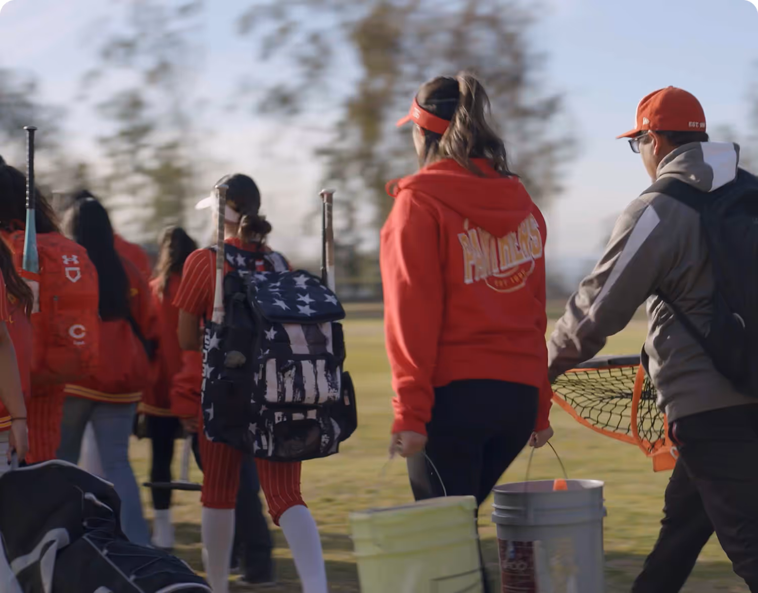 Softball team walking on a field carrying gear bags and buckets, with a coach in a red sweatshirt and cap.