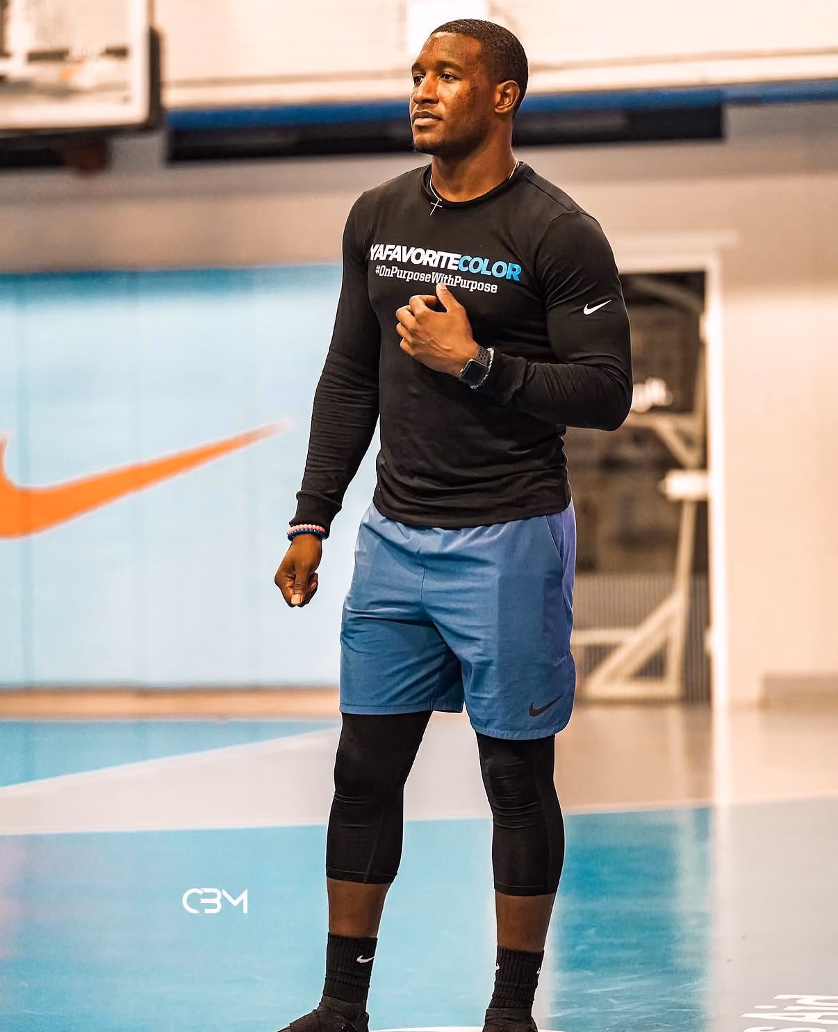 Man in black long sleeve shirt and blue shorts standing on a basketball court with Nike logos.
