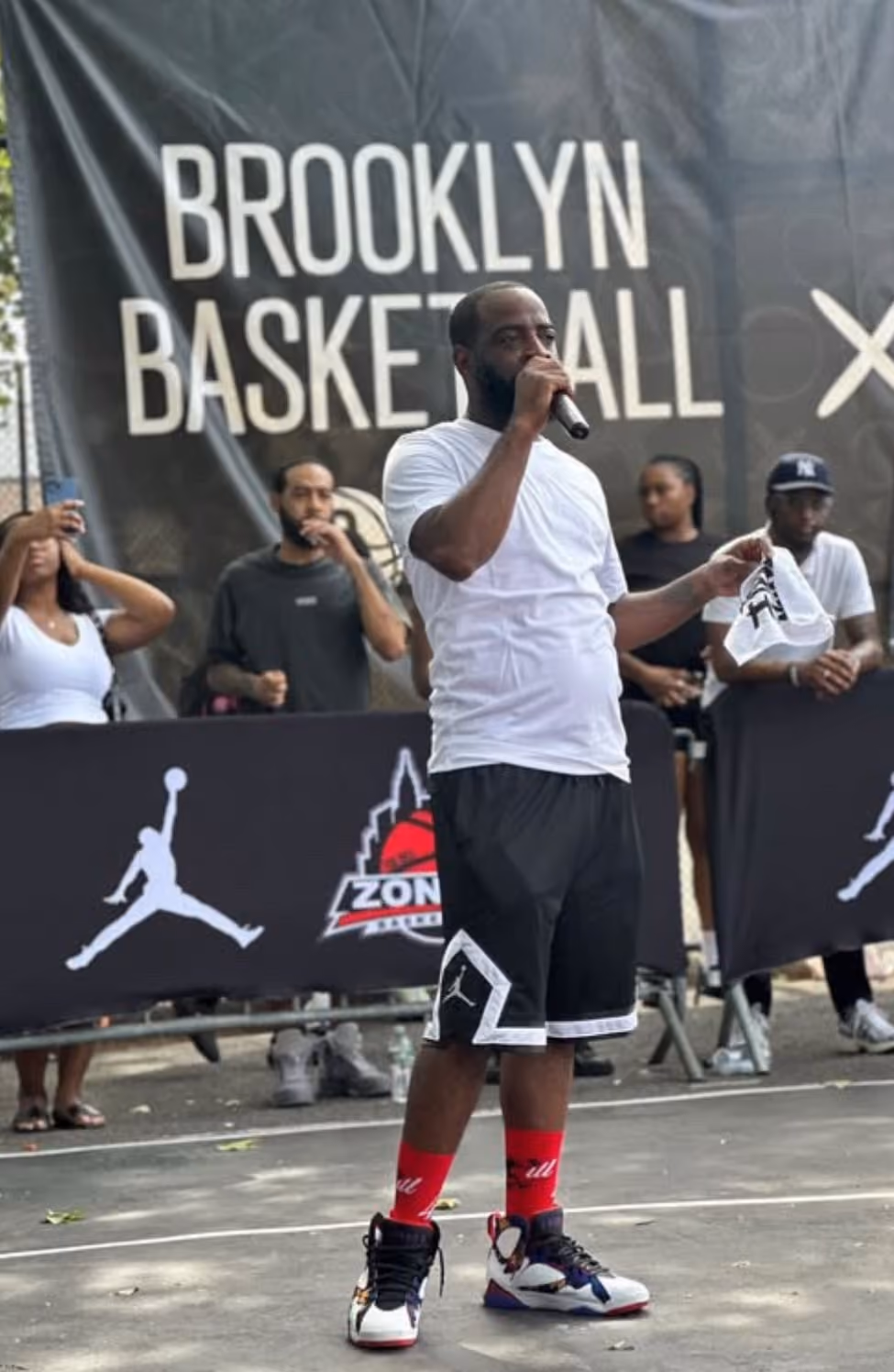 Man in white shirt speaking into microphone at Brooklyn Basketball event outdoors.