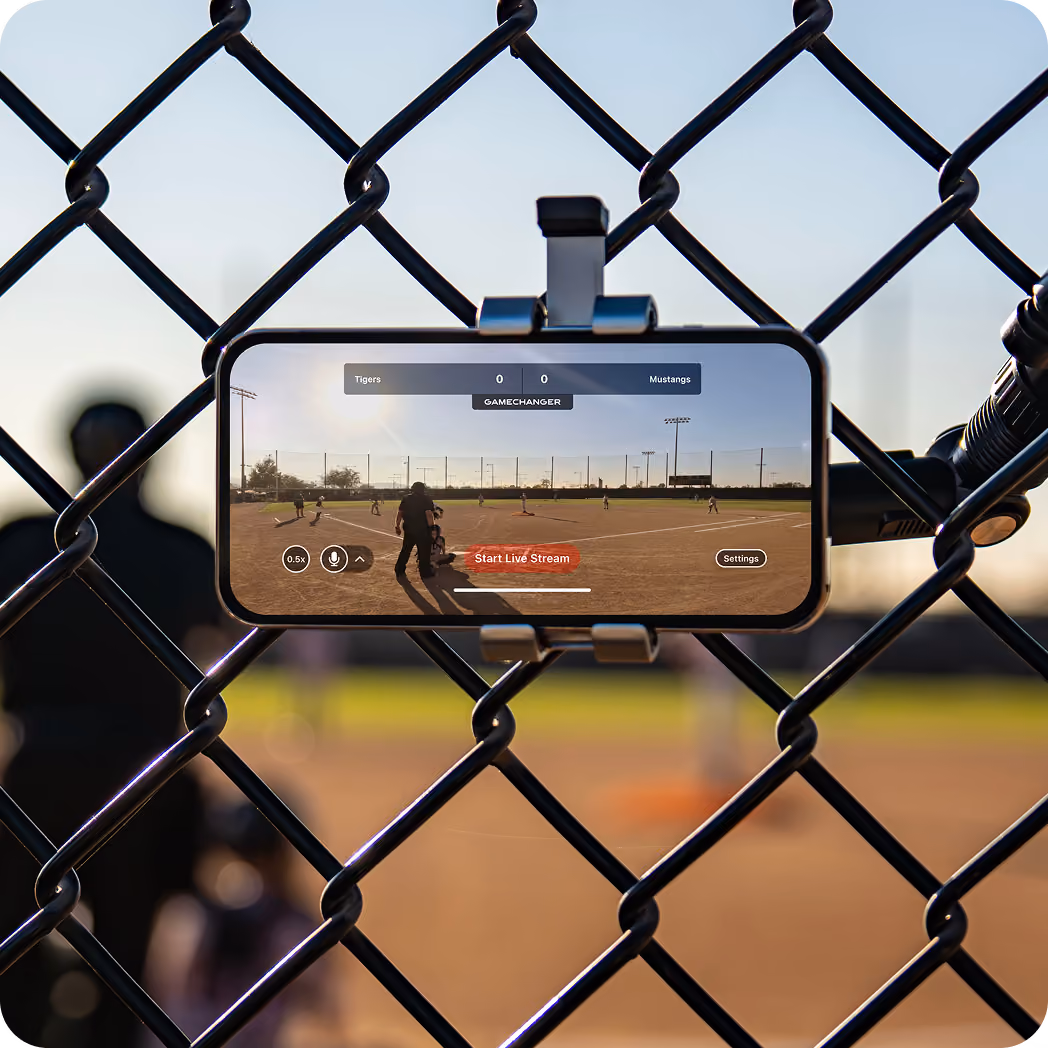 Smartphone mounted on chain-link fence live streaming a youth baseball game between Tigers and Mustangs.