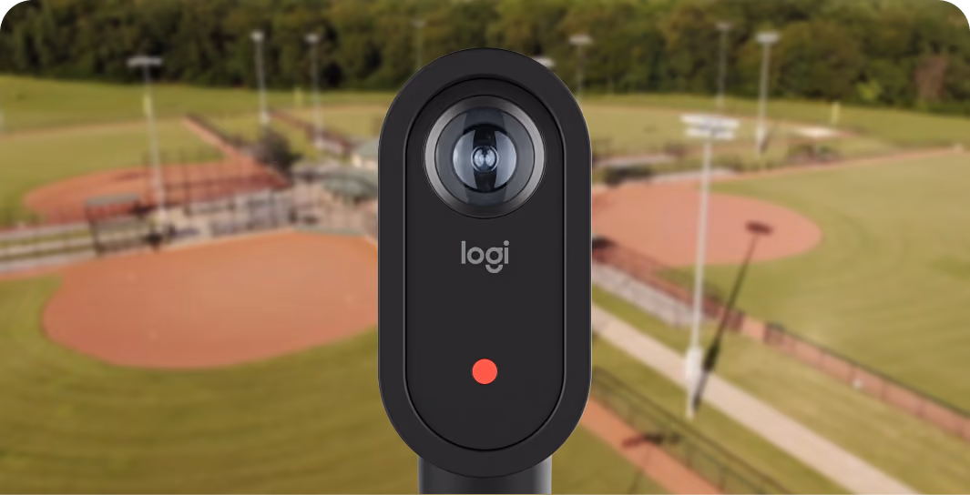 Black Logitech webcam with a red indicator light in front of a blurred baseball field background.