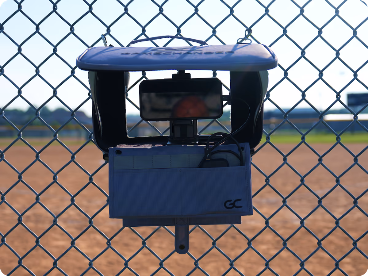 Mobile phone mounted on a device attached to a chain-link fence overlooking a baseball field.