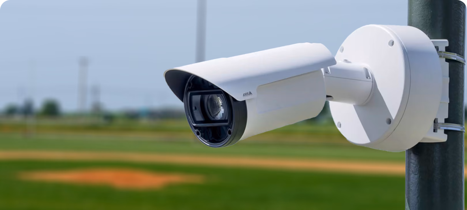White outdoor security camera mounted on a pole with a blurred grassy field background.