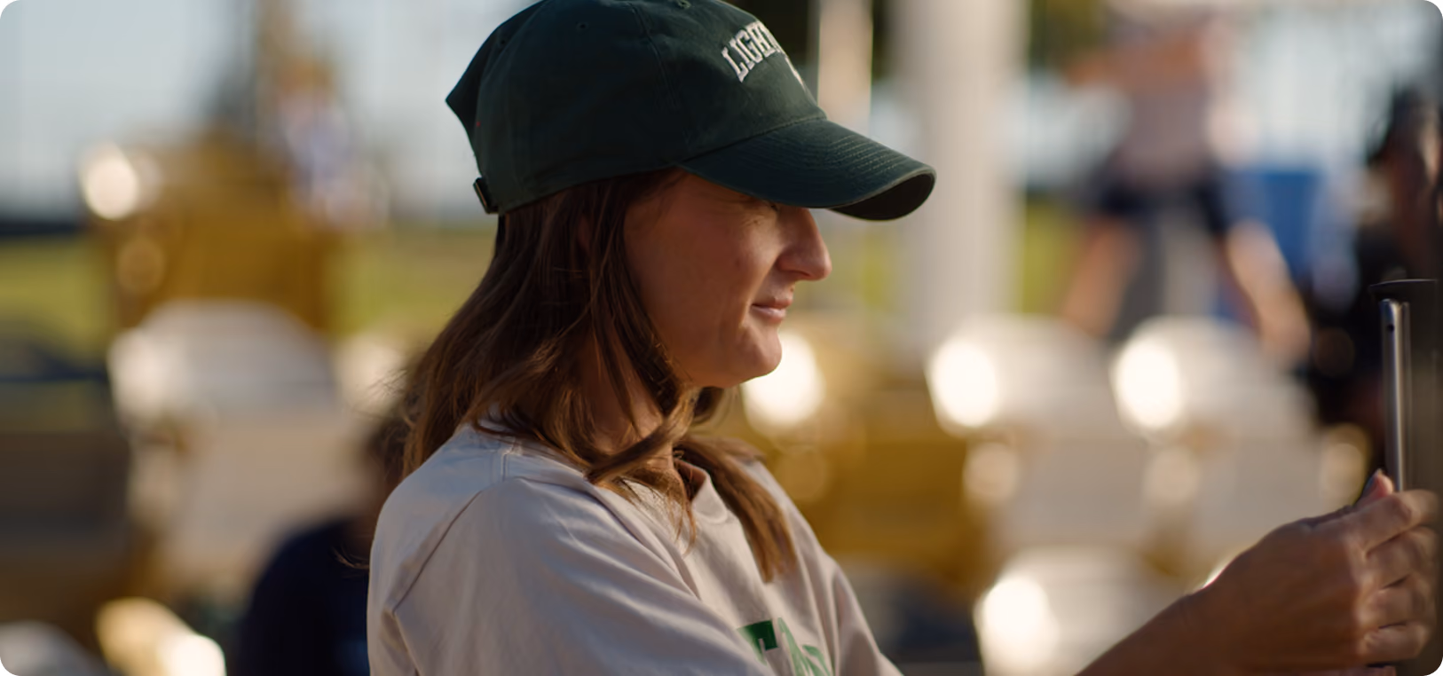 Profile of a woman wearing a black cap and light shirt holding a phone in a sunny outdoor setting.