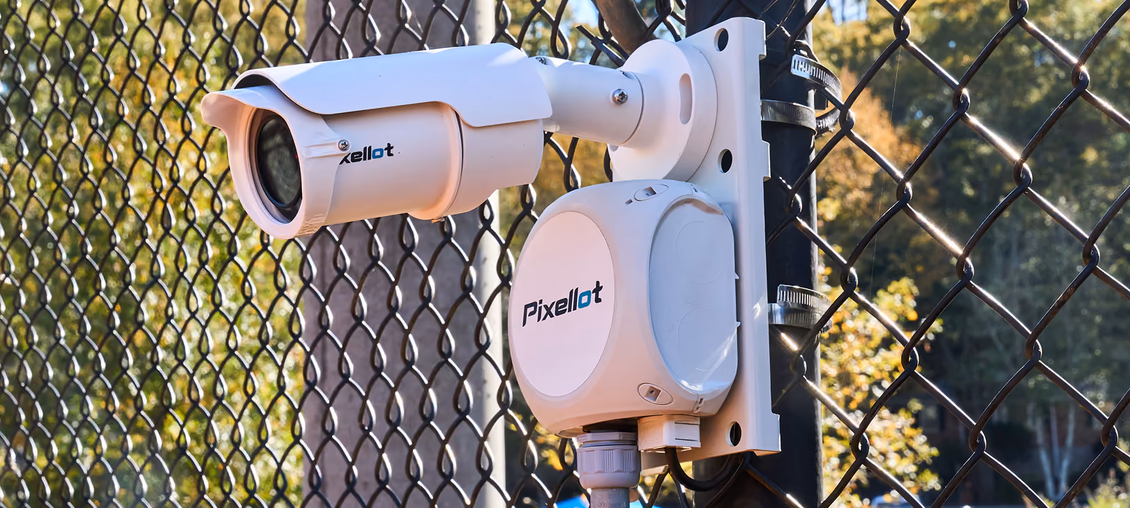 White Pixellot security camera mounted on a metal fence outdoors with trees in background.