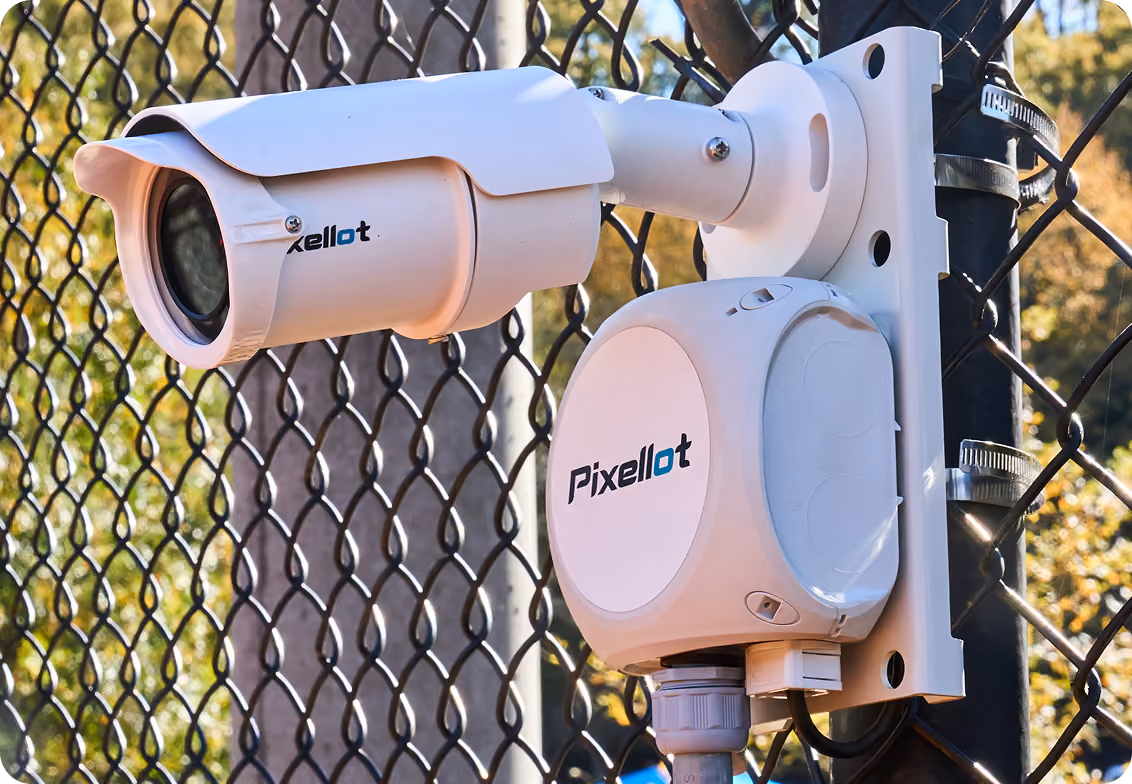 White Pixellot security camera mounted on metal bracket attached to a chain-link fence.