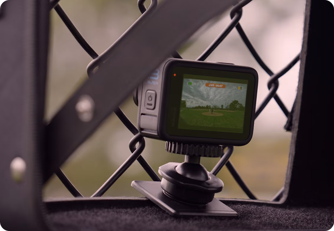 GoPro camera mounted on a metal surface recording a baseball game through a chain-link fence.