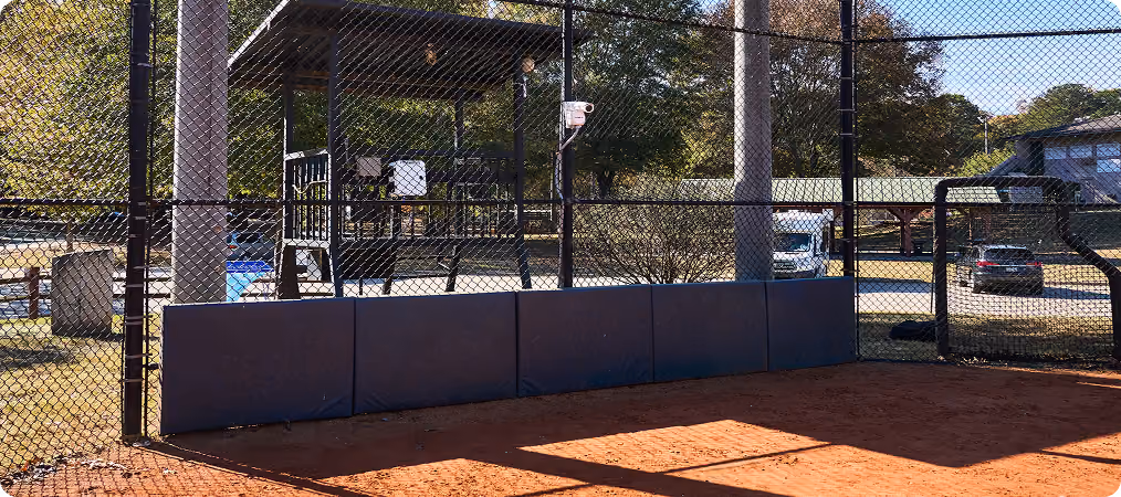 Empty baseball or softball batting cage with padded lower fence and a small covered seating area behind it.